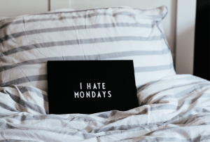 A black pillow on a bed that says I hate Mondays in white letters