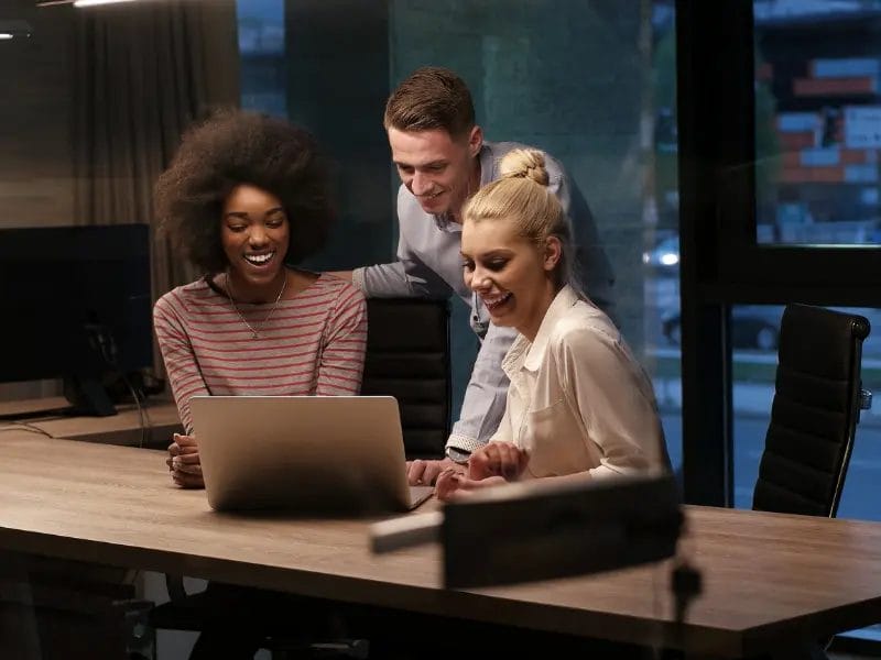 een groep mensen zit aan een tafel en kijkt naar een laptop.