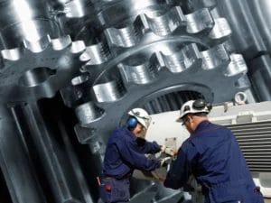 two men working on a machine with a large gear in the background