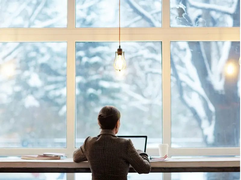 a woman is sitting at a desk in front of a window using a laptop .