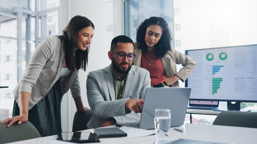 three professionals looking at a laptop with another screen with graphs at the back
